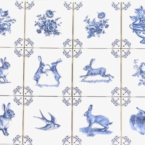 May include: A collection of twelve square ceramic tiles with a white background and blue illustrations. The tiles feature floral designs, rabbits, birds, and a fruit bowl. Each tile has a decorative border in the corners.