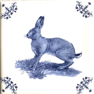 May include: A square ceramic tile featuring a blue ink-style illustration of a hare standing in grass. The design includes intricate blue floral corner accents on a white background, creating a detailed and classic decorative visual element.