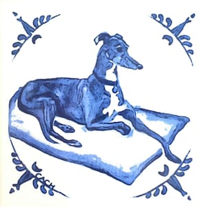 May include: A blue and white decorative tile featuring a greyhound dog resting on a pillow. The dog is painted in shades of blue with white accents. The tile is framed by a decorative border with floral elements.