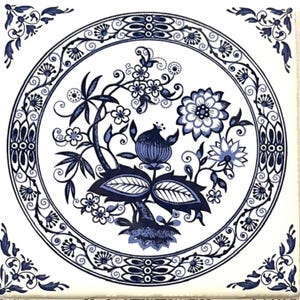 May include: A square ceramic tile featuring a white background with a dark blue circular border containing intricate floral and botanical patterns. The center displays a stylized plant motif with leaves, blossoms, and a central bulbous fruit.