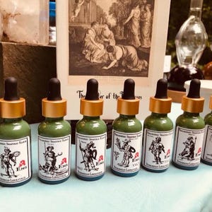 May include: Seven small green glass bottles with gold caps and labels. Each bottle has a different image of a person in a historical costume. The labels read "Alchemical Savaric Elixir" and "Ens" with a small red symbol.