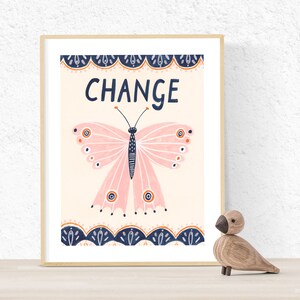 May include: A framed print featuring a pink butterfly with the word "CHANGE" in navy blue. The butterfly has orange and navy blue accents. The print is in a light wood frame, and a wooden bird figurine sits in front of it.