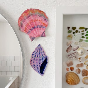 May include: Two seashell-shaped wall art pieces. One is a pink scallop shell with rainbow stripes. The other is a blue and pink conch shell. A framed display of sea glass and shells is visible.