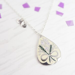 Puede incluir: Collar plateado con un colgante en forma de lágrima con un diseño floral en crema, verde y morado. El colgante tiene un detalle de lazo. Dos pequeños dijes redondos con las letras "N" y "O" cuelgan cerca del cierre.