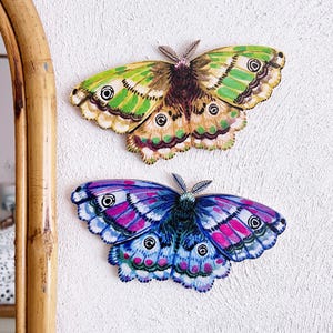 May include: Two decorative butterfly wall decals. The top butterfly is green, yellow, and brown with black accents. The bottom butterfly is blue, pink, and purple with black accents.