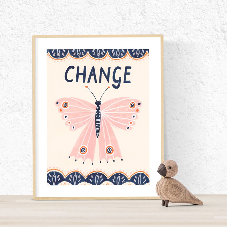 May include: A framed print featuring a pink butterfly with the word "CHANGE" in navy blue. The butterfly has orange and navy blue accents. The print is in a light wood frame, and a wooden bird figurine sits in front of it.