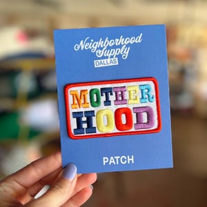 May include: A colorful embroidered patch with the word "MOTHERHOOD" in various colors, set against a white background with a red border. The patch is on a blue card that reads "Neighborhood Supply DALLAS" and "PATCH".