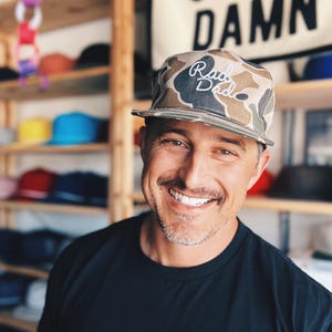 May include: A camouflage print baseball cap with the text "Rad Dad" in white script. The cap is worn by a smiling person in a black shirt. Shelves of hats are visible in the background, along with a sign that says "GIVE A DAMN".