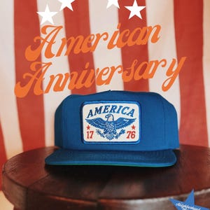 May include: A blue baseball cap with a rectangular patch on the front. The patch features a blue eagle graphic, the word AMERICA in blue text, and the numbers 17 and 76 in red on either side of the eagle. The cap has a flat brim and a structured crown.