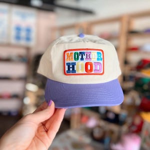 May include: A cream-colored baseball cap with a purple brim and a matching button on top. The cap features the word "MOTHERHOOD" embroidered in colorful letters within a rectangular patch.