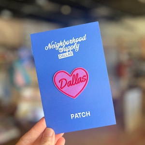 Puede incluir: Una tarjeta azul con el texto blanco "Neighborhood Supply DALLAS". Un parche rosa en forma de corazón con la palabra "Dallas" está centrado en la tarjeta. La palabra "PATCH" está impresa debajo del corazón.