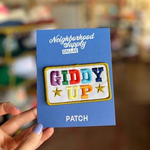 May include: Embroidered patch with the words "GIDDY UP" in colorful letters, outlined in gold, and two gold stars. The patch is on a blue card with "Neighborhood Supply DALLAS" printed at the top and "PATCH" at the bottom.