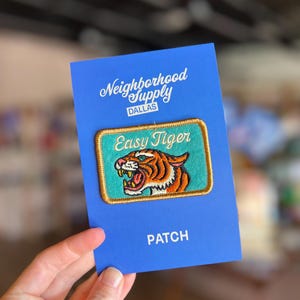 May include: A blue card with the text "Neighborhood Supply DALLAS" and "PATCH". The card holds a rectangular patch with a tiger design and the words "Easy Tiger". The tiger is orange, brown, and white.