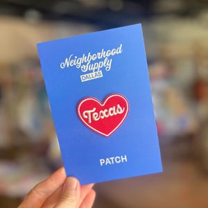 May include: A blue card with the text "Neighborhood Supply Dallas" and "PATCH". A red heart-shaped patch with the word "Texas" in white script is centered on the card. The patch has a white border.