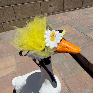Può includere: Una statua di oca bianca con becco arancione e un copricapo di tulle giallo. Il copricapo è adornato da una margherita bianca e una piccola ape, e un nastro nero. L'oca è posizionata su una superficie di mattoni.