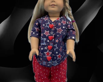 May include: A doll wearing a patriotic outfit. The ensemble includes a navy blue shirt with red and white stars, red heart-shaped buttons, and matching red shorts with white polka dots. The doll also wears a red hat with white polka dots and matching shoes.