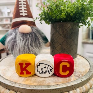 May include: Three wooden blocks with the letters K, C, and a heart with the word "chiefs" inside. The blocks are painted yellow, white, and red with the letters in red.