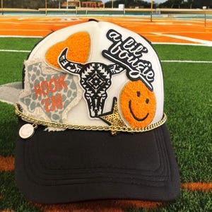 Könnte beinhalten: Eine schwarz-weiße Trucker-Kappe mit weißer Basis und schwarzem Schirm. Die Kappe hat mehrere Patches, darunter ein Herz, einen Totenkopf, ein Smiley und ein Patch mit der Aufschrift "a lil' bougie". Die Kappe hat auch eine goldene Kette mit einem Anhänger.