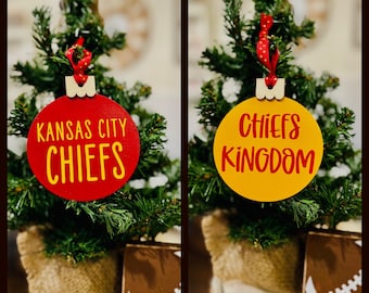 Op de afbeelding: Twee kerstversieringen met een Kansas City Chiefs-thema. Een versiering is rood met de tekst "Kansas City Chiefs" in geel. De andere versiering is geel met de tekst "Chiefs Kingdom" in rood.
