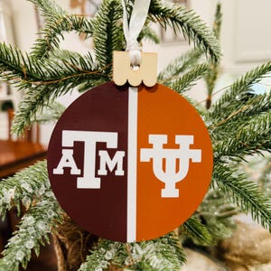 May include: A round, wooden Christmas ornament with a maroon and orange background. The ornament is divided in half, with the left side featuring the letters "A" and "M" in white, and the right side featuring the letter "U" in white. The ornament is hanging from a white ribbon on a green Christmas tree.