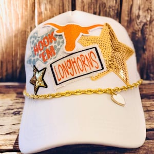 Könnte beinhalten: Weiße Baseballkappe mit goldener Kette und Herzanhänger. Die Kappe hat einen goldfarbenen Paillettenstern, ein orangefarbenes Longhorn-Logo und die Worte "Hook 'Em Longhorns" in Orange und Schwarz.