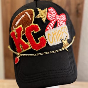 May include: Black trucker hat with a red and gold "KC Chiefs" patch, a red bow, a gold star, and a brown football patch. A gold chain with a red heart charm hangs from the hat.