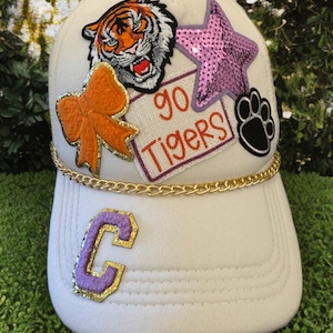 May include: White baseball cap adorned with patches including a tiger, star, paw print, and bow. The cap features the text "GO TIGERS" and a gold chain detail. A purple and gold letter "C" is on the front.