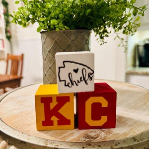 May include: Three wooden blocks spelling "KC" with a white block on top that says "chiefs" with a heart in the state of Missouri outline. The blocks are on a wooden table with a beaded edge.