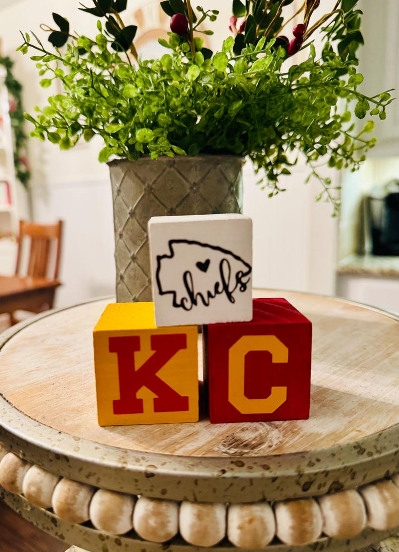 May include: Three wooden blocks spelling "KC" with a white block on top that says "chiefs" with a heart in the state of Missouri outline. The blocks are on a wooden table with a beaded edge.