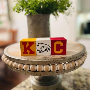 May include: Three wooden blocks spelling "KC" with a heart and the word "chiefs" on the middle block. The blocks are painted in red, yellow, and white. The blocks are sitting on a wooden cake stand with a beaded edge.