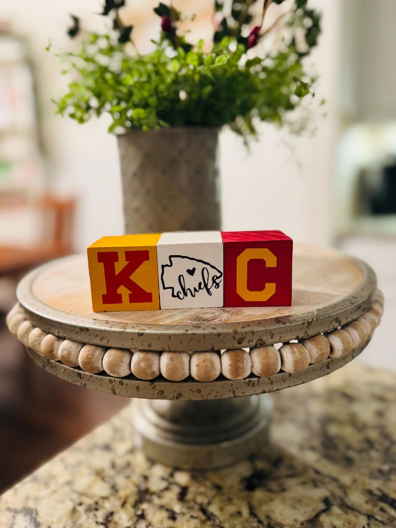 May include: Three wooden blocks spelling "KC" with a heart and the word "chiefs" on the middle block. The blocks are painted in red, yellow, and white. The blocks are sitting on a wooden cake stand with a beaded edge.