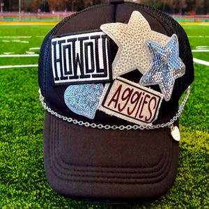 May include: Black trucker hat with a silver chain and a heart charm. The hat has several patches, including a white star, a silver star, a blue heart, and a patch that says "Howdy Aggies".