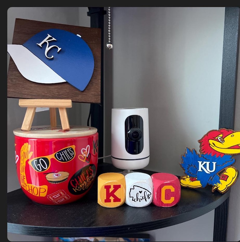 May include: A shelf with a red and white mug with the words "Go Chiefs" and "KC Chiefs Kingdom" on it. There are also three wooden blocks that spell out "KC" and a wooden cutout of a blue and red bird with the letters "KU" on it.