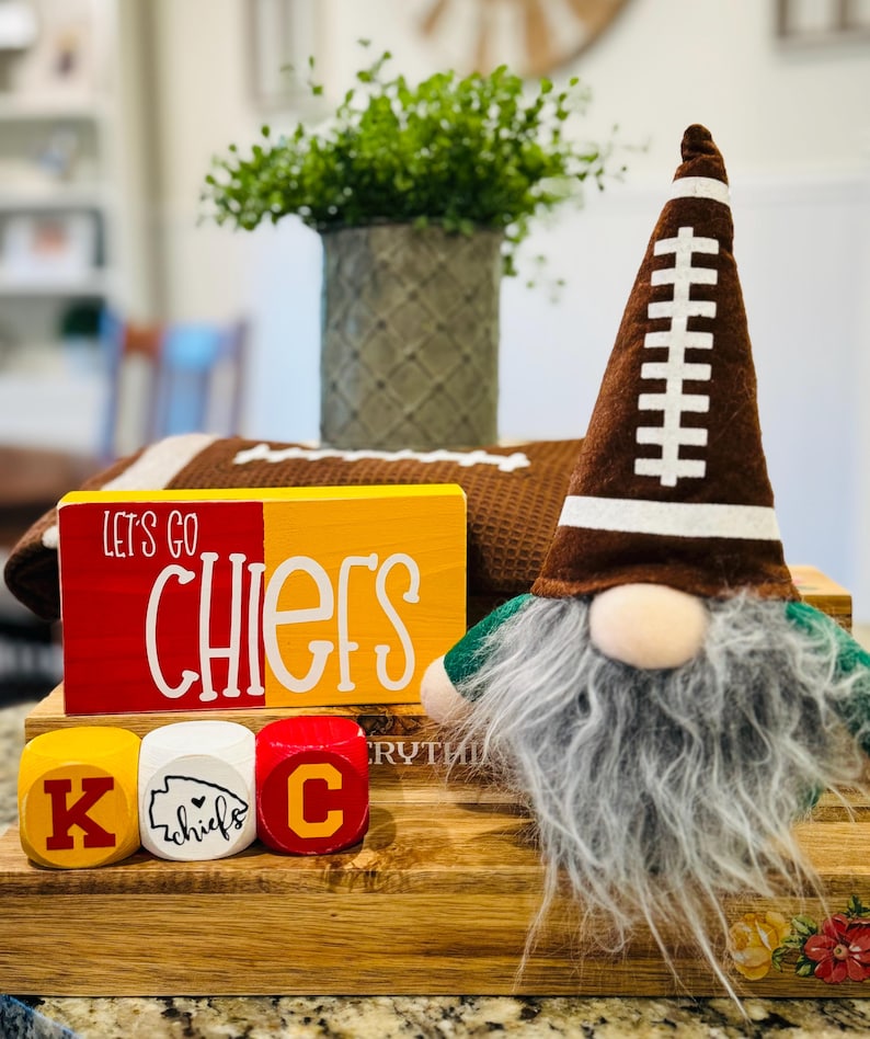 May include: A wooden sign with the words "Let's Go Chiefs" in red and yellow paint.  The sign is sitting on a stack of wooden blocks that spell out "K.C."  A football-themed gnome with a brown hat and a white beard is sitting in front of the sign.