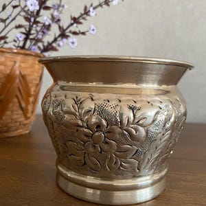May include: A silver planter with an embossed floral design. The planter is round and has a wide base.