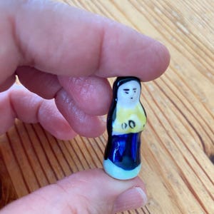 May include: A small, hand-painted ceramic figurine shaped like a person with a black head covering, a yellow upper garment, and a dark blue lower garment. The figure has a simple painted face with black eyes and a small mouth, featuring a glossy finish.