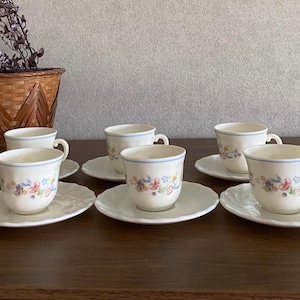 May include: A set of three off-white ceramic teacups and matching saucers, each featuring a delicate floral pattern in pink, blue, and green with a thin blue rim. The saucers have a scalloped edge design, and the cups have a rounded shape.