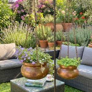 Op de afbeelding: Twee gehamerde koperen hangplantenbakken gevuld met groen en bloemen hangen aan kettingen. De plantenbakken staan in een tuin. De plantenbakken zijn rond met een gehamerde textuur.