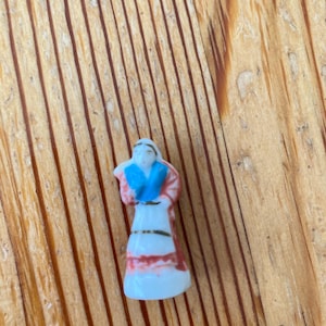 May include: A small porcelain figurine of a person standing upright. The figure wears a white garment with a blue top section, a red shawl or wrap draped over the shoulders, and thin gold-colored horizontal bands around the waist and skirt area.