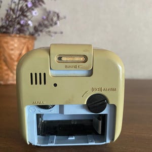 K&ouml;nnte beinhalten: Ein beiger und blauer Vintage-Wecker mit einem schwarzen Alarmknopf und einem Schiebeschalter mit der Aufschrift "(SNZ)". Die Uhr hat ein kleines Fenster, das die Zeit anzeigt, und die W&ouml;rter "JAPAN" und "ALARM" sind auf der Vorderseite aufgedruckt.
