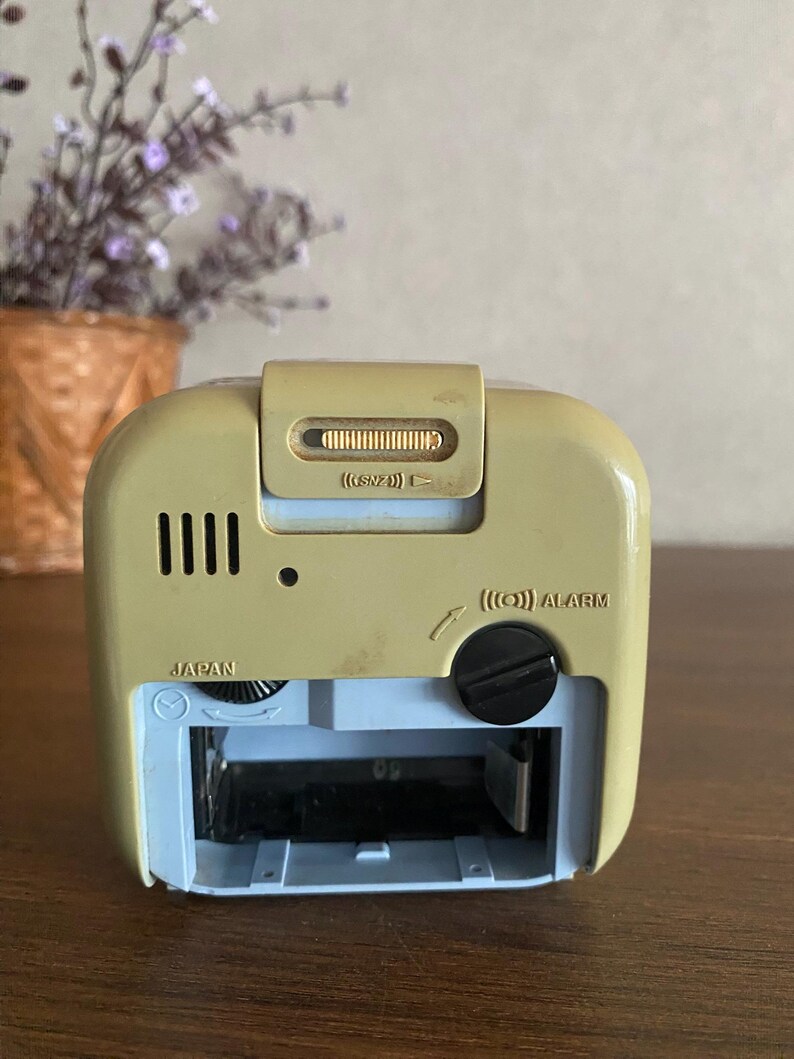 K&ouml;nnte beinhalten: Ein beiger und blauer Vintage-Wecker mit einem schwarzen Alarmknopf und einem Schiebeschalter mit der Aufschrift "(SNZ)". Die Uhr hat ein kleines Fenster, das die Zeit anzeigt, und die W&ouml;rter "JAPAN" und "ALARM" sind auf der Vorderseite aufgedruckt.