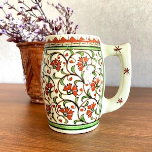 Könnte beinhalten: Cremefarbene Keramiktasse mit Henkel, verziert mit roten Blumenmustern und grünen Ranken. Der Rand hat ein rot-braunes Design. Der Griff hat sternförmige Akzente. Ein dekoratives Stück für die Küche oder das Zuhause.