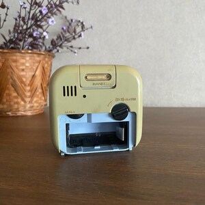 K&ouml;nnte beinhalten: R&uuml;ckansicht eines beigen Weckers mit einem Batteriefach mit der Aufschrift "Japan". Die Uhr hat einen schwarzen Knopf mit der Aufschrift "(L) ALARM" und einen Schieberegler mit der Aufschrift "(L)  SOUND  (H)".