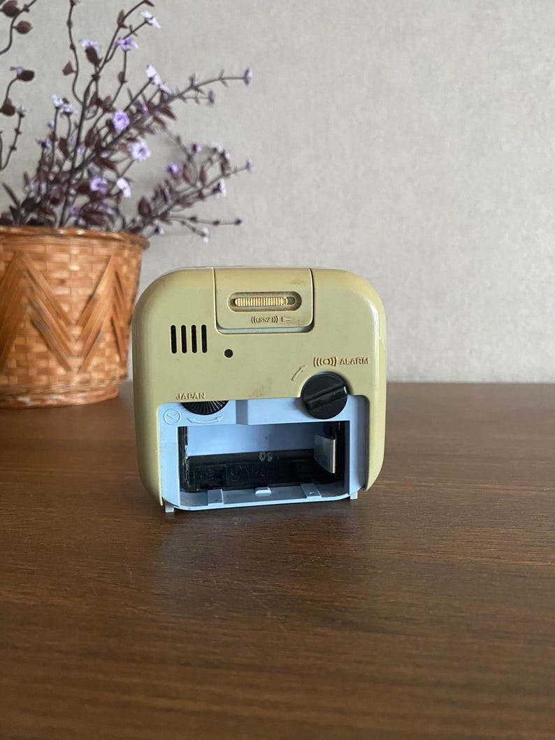 K&ouml;nnte beinhalten: R&uuml;ckansicht eines beigen Weckers mit einem Batteriefach mit der Aufschrift "Japan". Die Uhr hat einen schwarzen Knopf mit der Aufschrift "(L) ALARM" und einen Schieberegler mit der Aufschrift "(L)  SOUND  (H)".