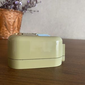 K&ouml;nnte beinhalten: Ein beiger Vintage-Wecker mit wei&szlig;em Zifferblatt und schwarzen Zeigern. Der Wecker steht auf einer Holzoberfl&auml;che.