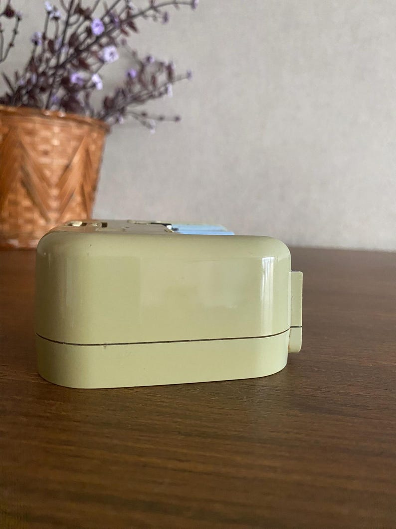 K&ouml;nnte beinhalten: Ein beiger Vintage-Wecker mit wei&szlig;em Zifferblatt und schwarzen Zeigern. Der Wecker steht auf einer Holzoberfl&auml;che.