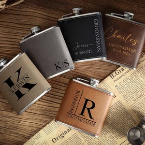 May include: An assortment of personalized flasks in various colors, including tan, gray, and brown, with silver tops. Each flask features custom text, such as names, initials, and the word "Groomsman". The flasks are arranged on a wooden surface.