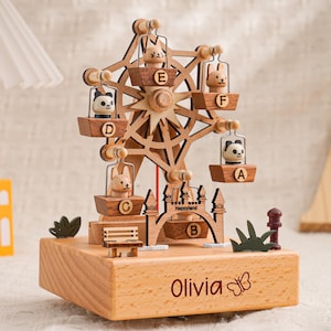 May include: A wooden Ferris wheel music box with six animal-shaped carriages. The base is engraved with the name "Olivia" and a butterfly. The Ferris wheel is made of light brown wood and features a castle and a bench.