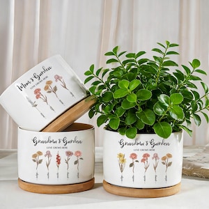 Puede incluir: Maceteros de cerámica blanca con platillos de madera. Las macetas están decoradas con ilustraciones florales y el texto "Mom's Garden" y "Grandma's Garden". Una maceta contiene una planta verde.