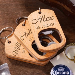 May include: Three rectangular wooden bottle openers with metal key rings. Each opener is engraved with a name and a date, including Alex 05.15.2026, Anthony, and William. The light-colored wood features a smooth finish and a central cutout.